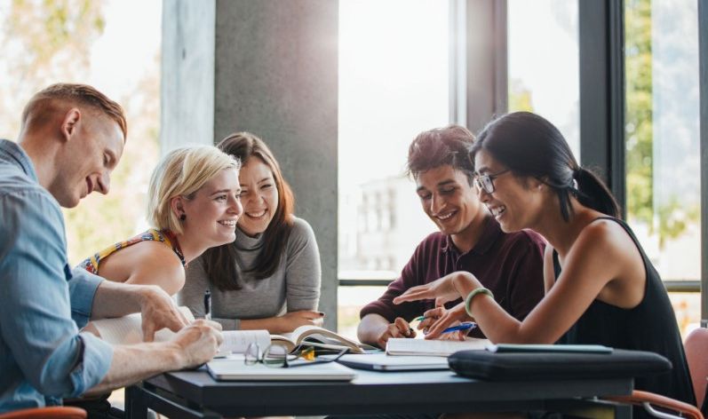 A group of five diverse young people sitting at a table by large windows with trees visible outside. They appear to be students or colleagues collaborating on a project. The group is engaged in conversation and looking at materials spread out on the table, including books and papers. Their expressions are cheerful and focused, suggesting a positive and productive atmosphere. The setting looks like a bright, modern study area or workspace.